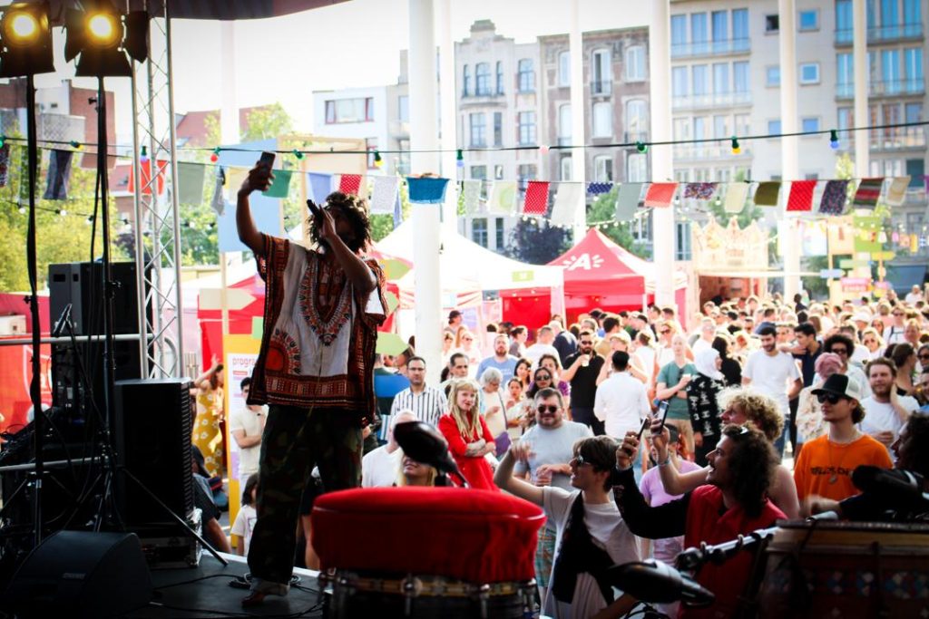 Person on stage taking a selfie with a crowd in the background at an outdoor festival. Colorful banners and buildings are visible.